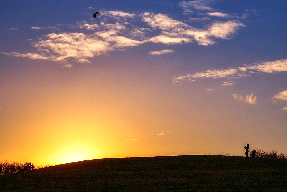 Glowing sun sinking behind a hill while a silhouetted person stands flying a kite, South Shields, Tyne and Wear, England