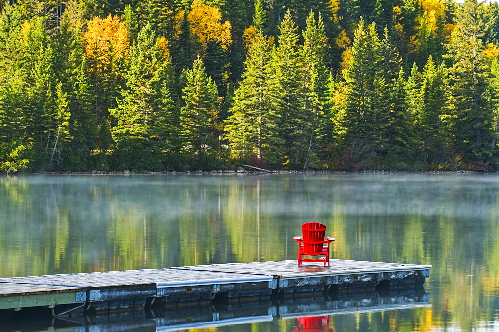 Muskoka chair on dock with autumn coloured foliage reflected in the tranquil lake water of Clear Lake, Riding Mountain National Park, Manitoba, Canada