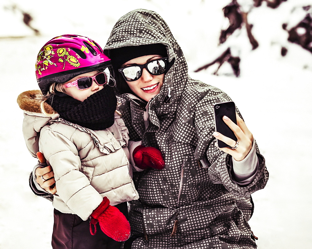 A Mother And Daughter Dressed In Winter Coats, Hats And Sunglasses Taking A Self-Portrait With A Smart Phone While Tobogganing on Vacation, Fairmont Hot Springs, British Columbia, Canada