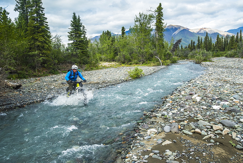 A man fat biking across a stream in Wrangell-St. Elias National Park and Preserve on a cloudy summer day in South-central Alaska, Alaska, United States of America