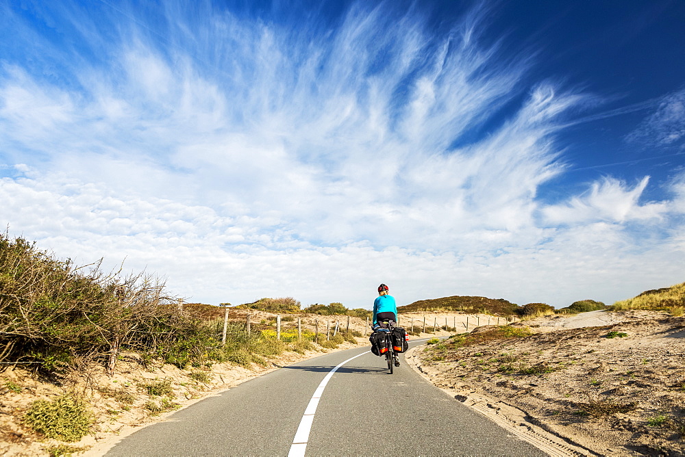 Female cyclist on paved bike pathway along rolling sand dunes with dramatic clouds and blue sky, South of Zandvoort, Netherlands