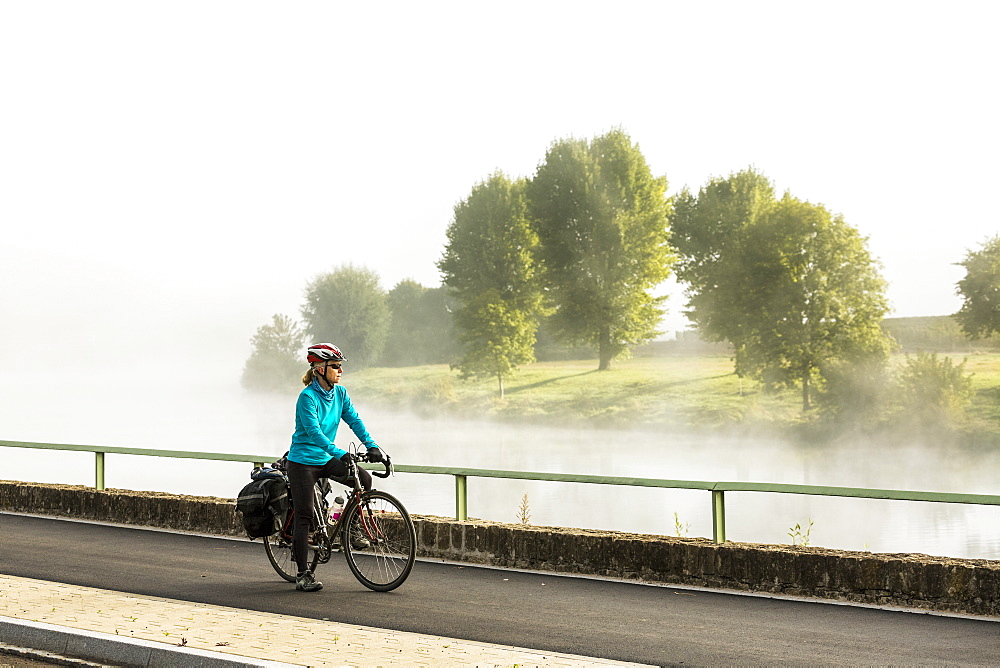 Female cyclist along river pathway with mist in the river valley looking over trees along river bank, North of Remich, Luxembourg