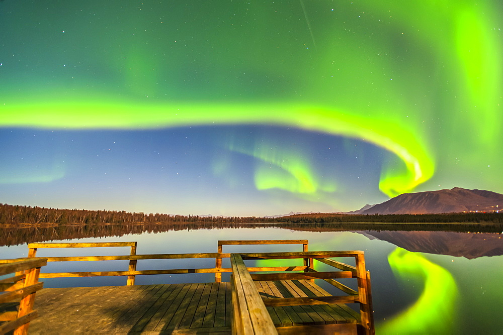 The Northern Lights are seen reflecting off Beach Lake on a clear, autumn night with a wooden dock in the foreground, Chugiak, Alaska, United States of America