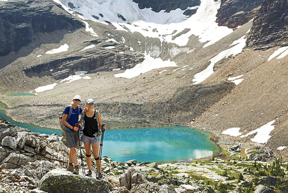 Two female hikers standing in a large rocky area with a colourful alpine lake and mountain cliffs with snow in the background, British Columbia, Canada