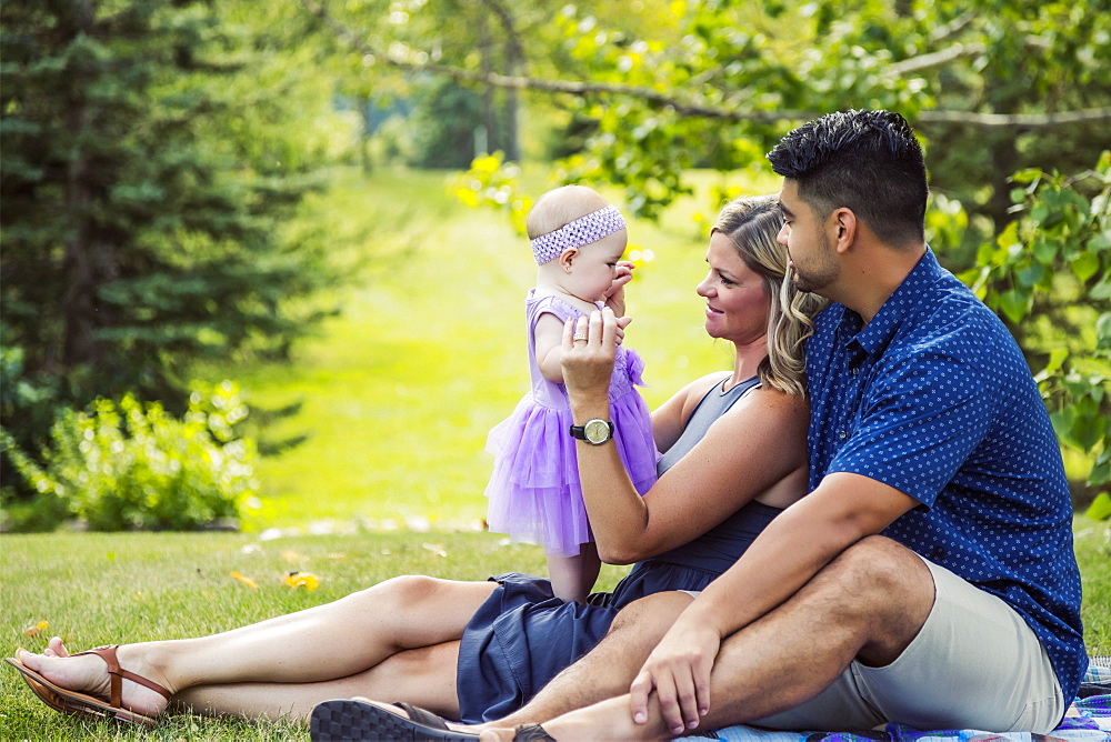 A mixed race husband and wife relax together with their baby girl on a blanket in a park on a warm autumn afternoon, Edmonton, Alberta, Canada