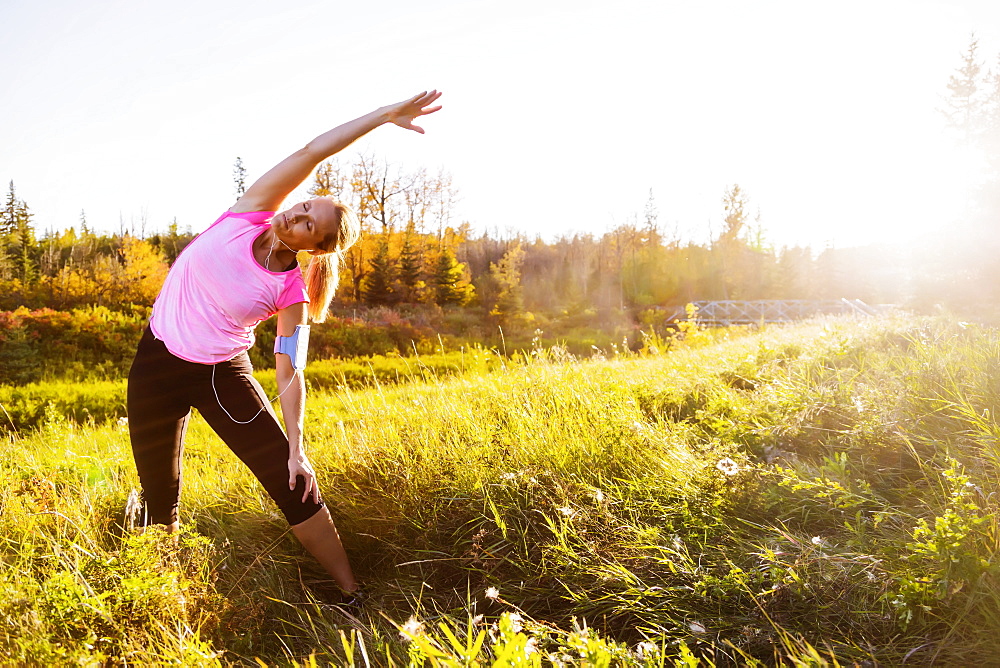 An attractive middle-aged woman wearing active wear and listening to music while pausing to do some stretch exercises during a run in a city park at sunset on a warm autumn evening, Edmonton, Alberta, Canada