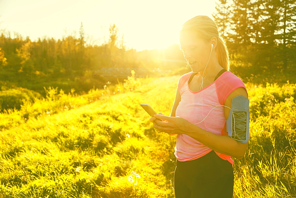 An attractive middle-aged woman wearing active wear pauses to text message while out running beside a creek in a city park on a warm fall evening and listening to music on her smart phone, Edmonton, Alberta, Canada