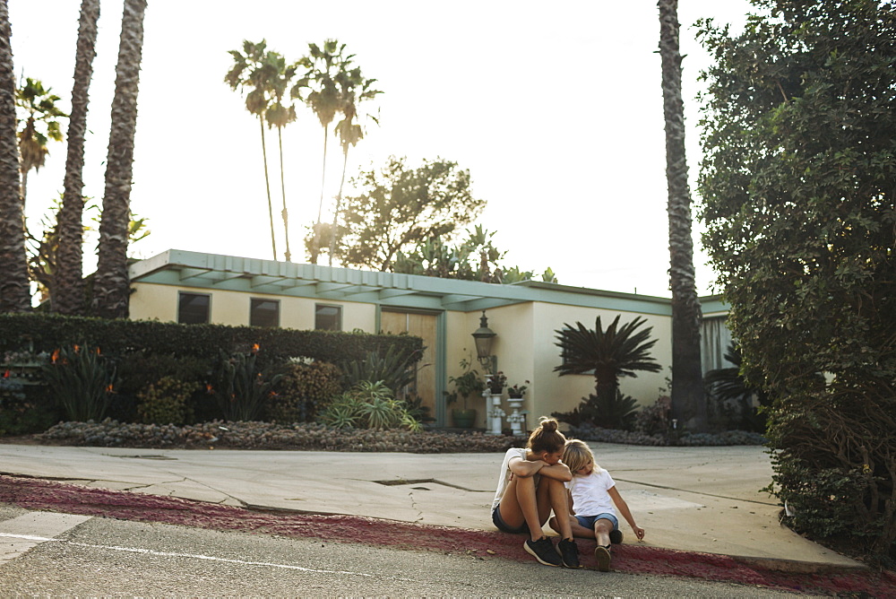 Two young girls sit on a street curb outside a house, Los Angeles, California, United States of America