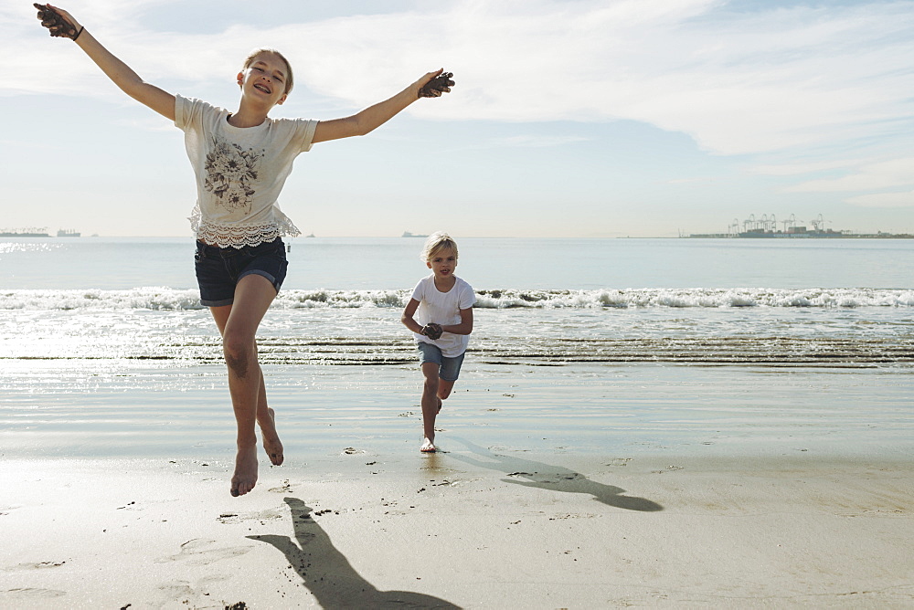 Two sisters running and playing on a beach, the oldest one in front making eye contact with the camera and she leaps into the air, Long Beach, California, United States of America