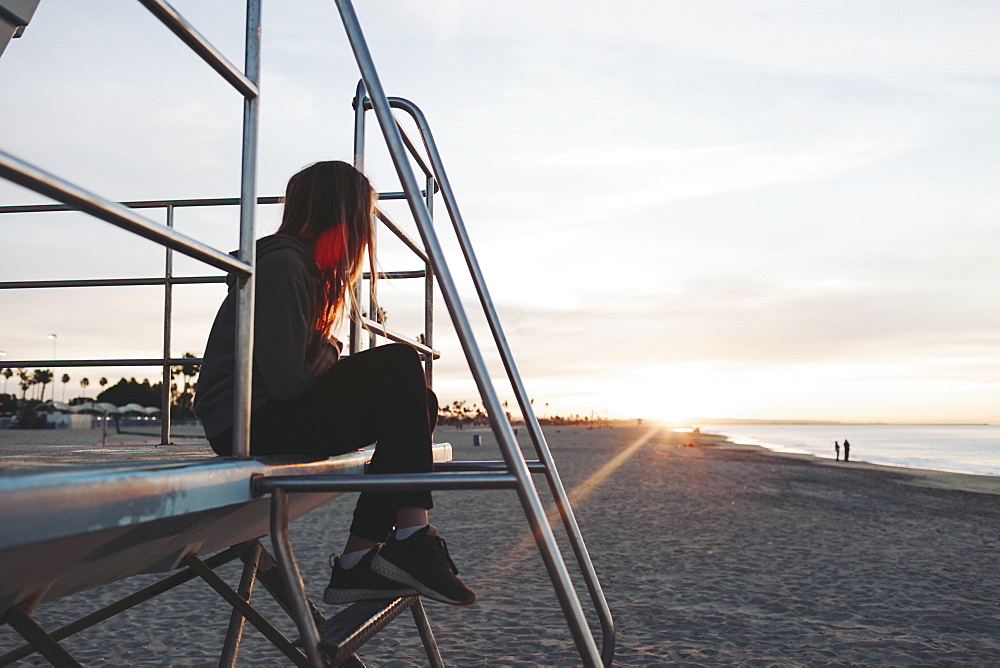 A girl sits on a lifeguard station at dawn looking out to the ocean, Long Beach, California, United States of America