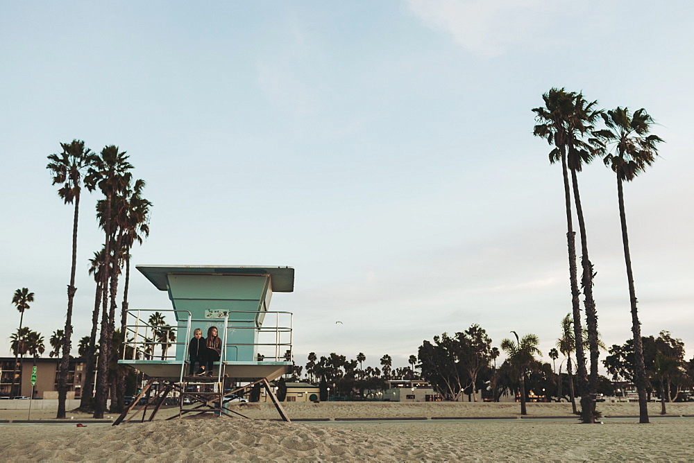 Two girls sit on a lifeguard station at dawn looking out to the ocean, Long Beach, California, United States of America