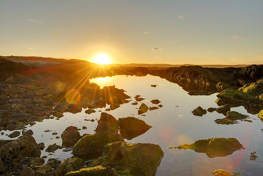 A golden-coloured sunset and sunburst hilights the rocks around an ocean-side pool of water, Chubut, Argentina