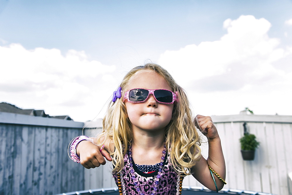 A young girl all dressed up with blond curly hair, sunglasses and jewelry standing on a trampoline in the backyard and showing off, Spruce Grove, Alberta, Canada