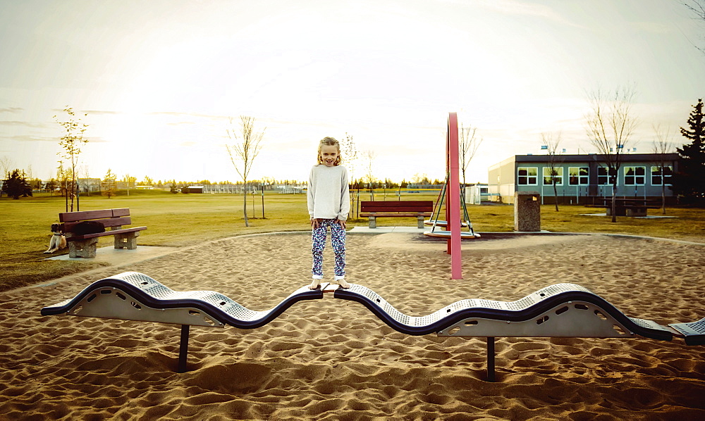 A young girl posing for the camera while standing on a bouncing balance beam in a playground on a warm autumn evening at sunset, Edmonton, Alberta, Canada