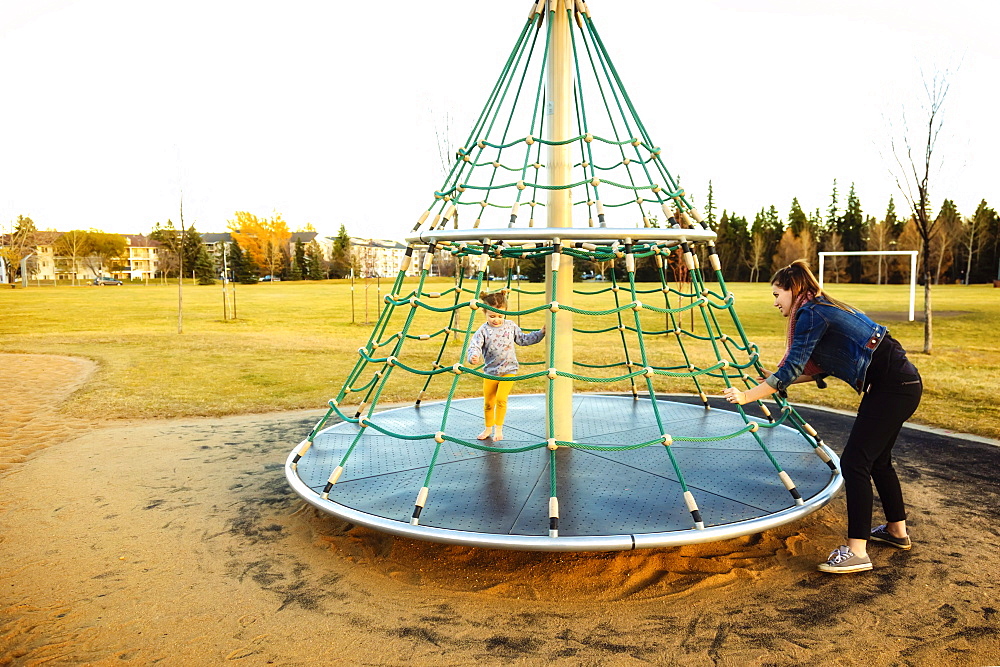 A young mom spinning her daughter while playing on a merry go round with a rope climber in a playground at sunset during a warm autumn evening, Edmonton, Alberta, Canada