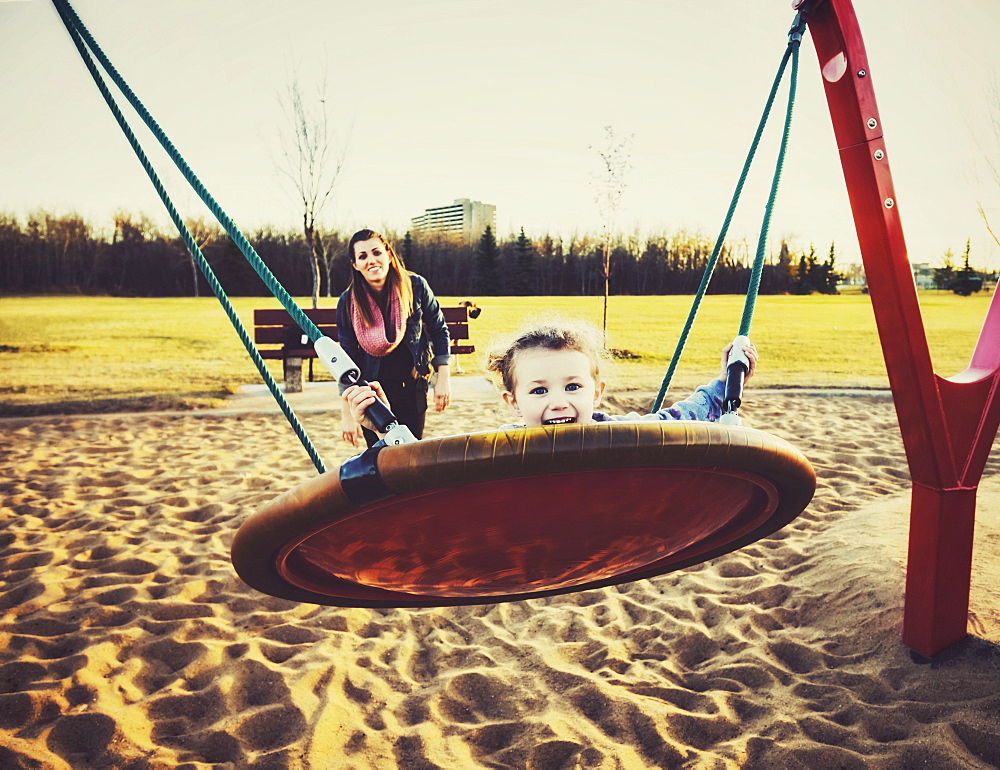 A young mom and her daughter playing on a saucer swing in a playground on a warm autumn evening, Edmonton, Alberta, Canada