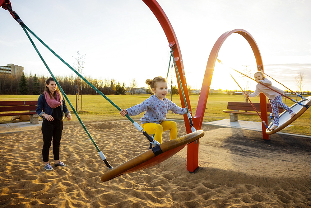 A young mom and her daughters playing on saucer swings in a playground on a warm autumn evening, Edmonton, Alberta, Canada