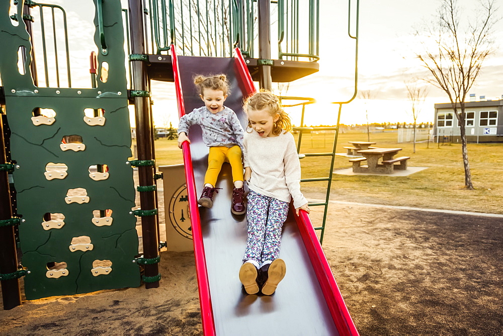 Two young sisters going down a slide in a playground on a warm autumn evening at sunset, Edmonton, Alberta, Canada