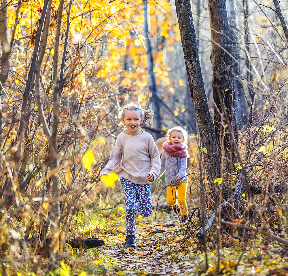 Two young girls who are sisters running through the woods in a city park on a warm fall evening, Edmonton, Alberta, Canada