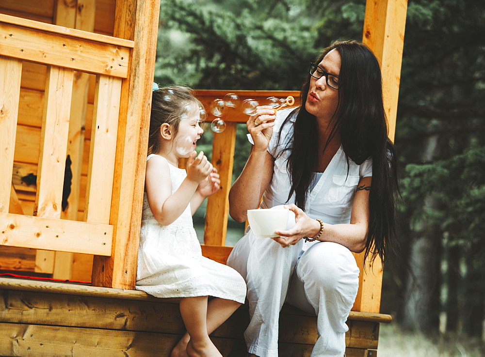 Mother and young daughter blowing bubbles on the steps of a wooden playhouse, Alberta, Canada