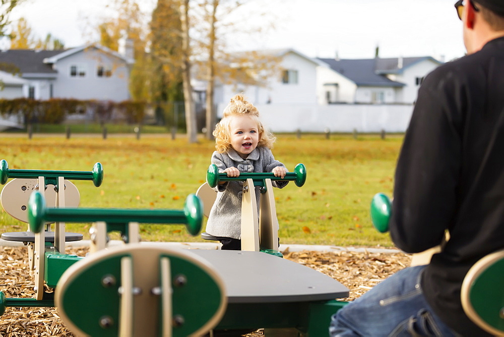 A cute young girl playing on a seesaw with her dad in a playground during the fall season, Spruce Grove, Alberta, Canada