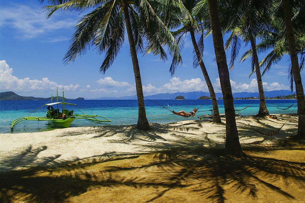 A woman lays on a hammock on a tropical beach with an outrigger canoe moored along the shore, Andaman Islands, India