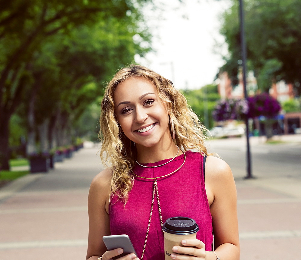 A young woman looks at the camera while walking down a street with a coffee near a university campus and texting on her smart phone, Edmonton, Alberta, Canada