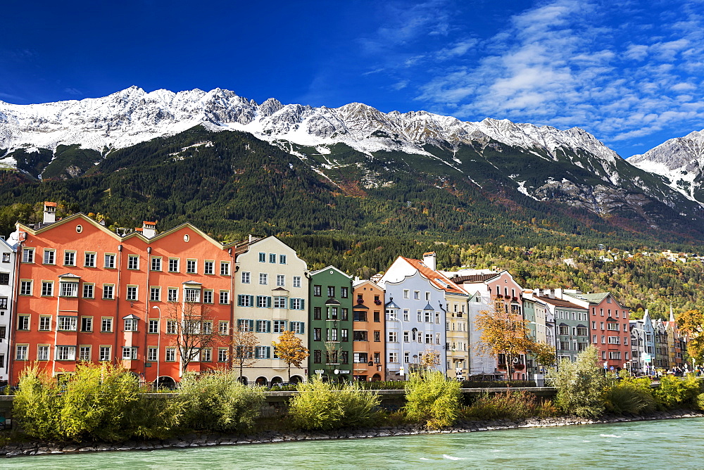 Colourful row of buildings along a riverbank with snow-covered mountain range in the background and blue sky, Innsbruck, Tyrol, Austria