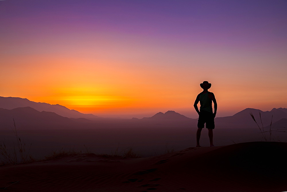 Silhouette of a man wearing a cowboy hat stands looking out at the glowing landscape and sky at sunset, Sossusvlei, Hardap Region, Namibia