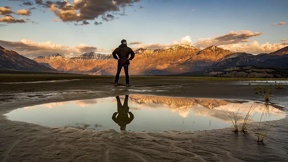 A man stands with his reflection in a pool of water looking out over the Saint Elias Mountains at sunset, Kluane National Park and Reserve, Destruction Bay, Yukon, Canada