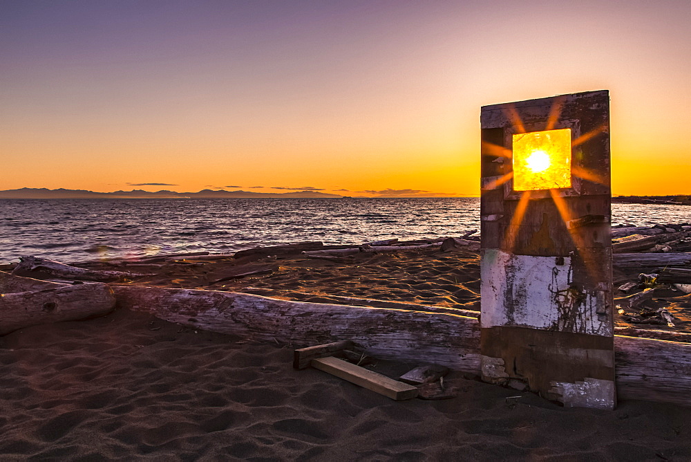 A sunburst shines bright through the window of a weathered, wooden door propped up on Iona beach, Richmond, British Columbia, Canada