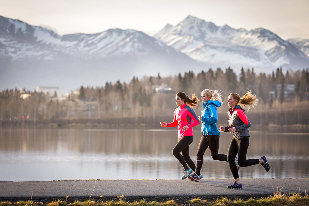 Three young women running on a trail along the water's edge with mountains in the distance, Anchorage, Alaska, United States of America