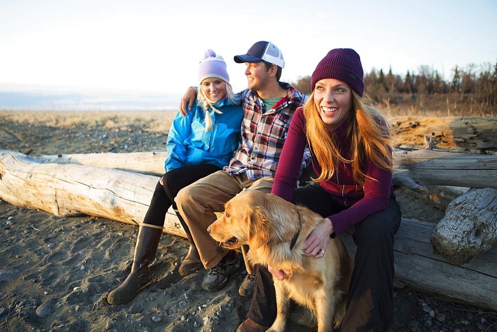 A young couple and a friend with a dog sit on a piece of driftwood on a beach looking out to the ocean at sunset, Anchorage, Alaska, United States of America