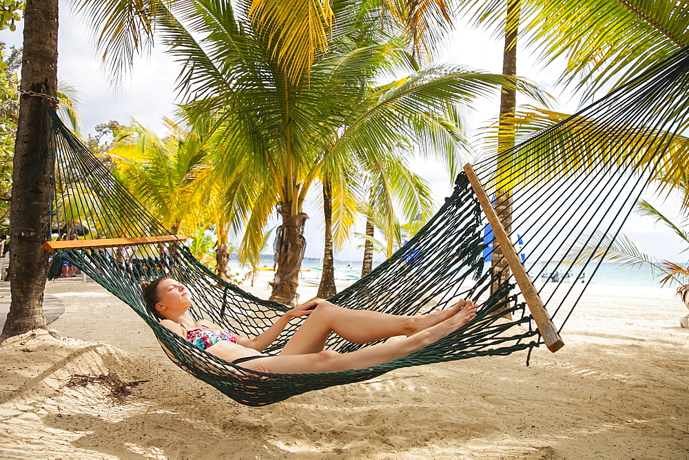 A young woman in a bikini lays in a hammock on a tropical beach with the ocean in the background, Negril, Jamaica