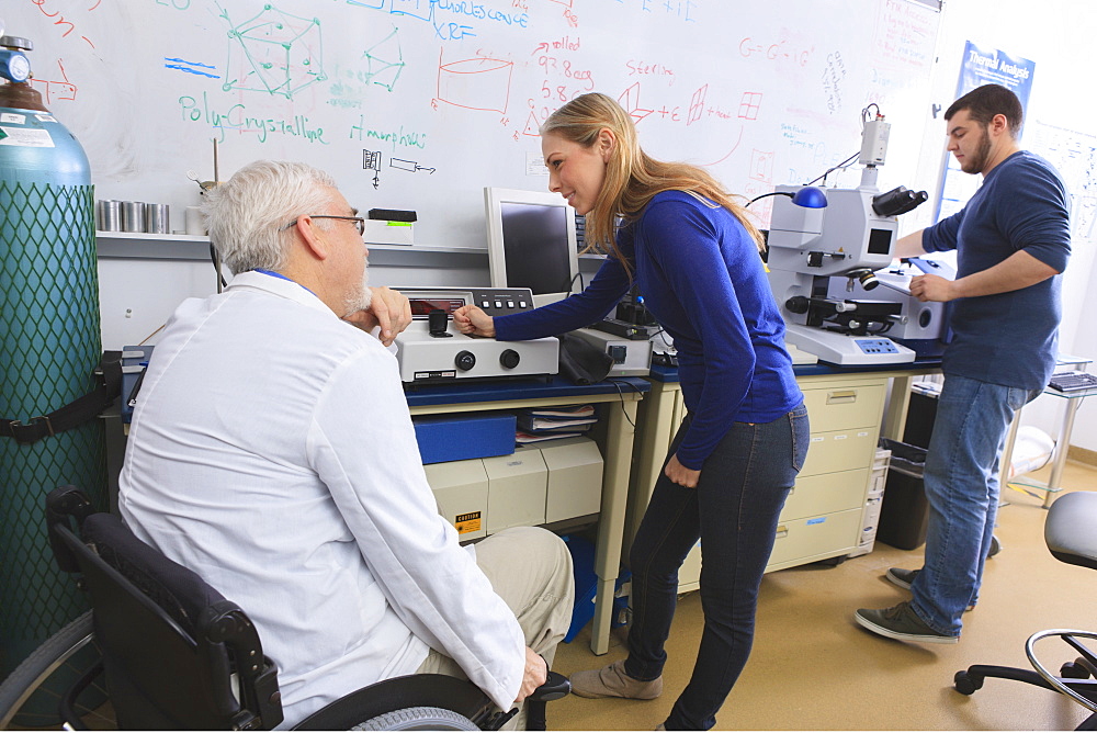 Professor with muscular dystrophy advising engineering students in chemistry laboratory working on x-ray fluorescence analyzers