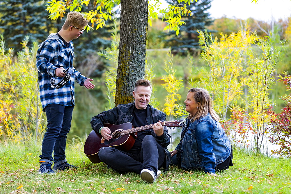 A young man with Down Syndrome playing a tambourine while his father plays a guitar and his mother sings along while enjoying each other's company in a city park on a warm fall evening: Edmonton, Alberta, Canada