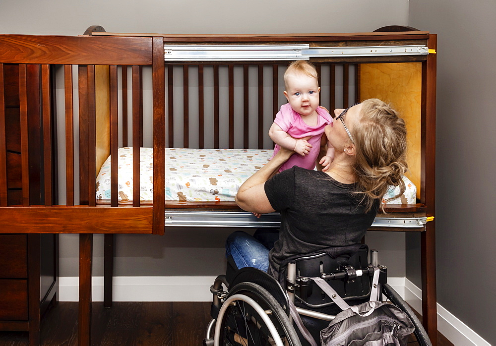 A paraplegic mother lifting a baby from a customized side-opening crib that allows her to put her baby down for a nap from her position in a wheelchair: Edmonton, Alberta, Canada