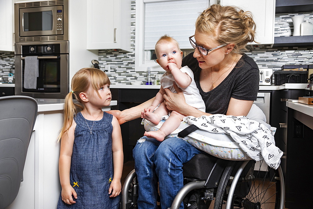 A paraplegic mom in a wheelchair talking with her daughter and holding her baby in her lap while working in her kitchen; Edmonton, Alberta, Canada