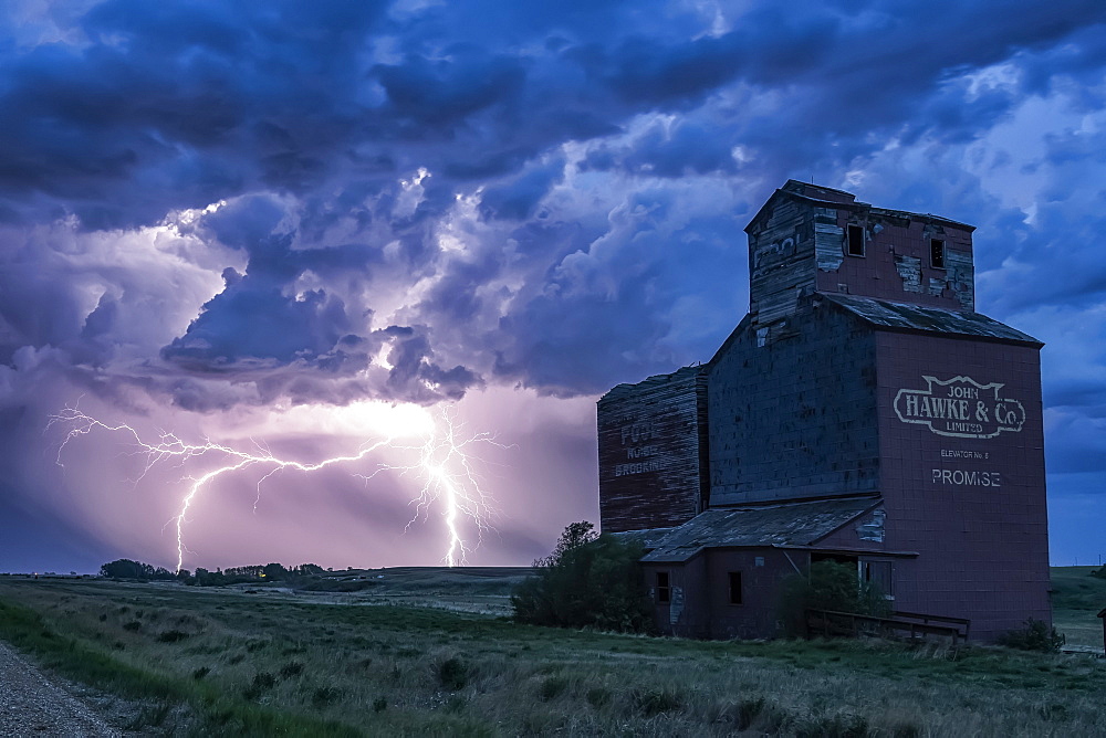 Grain elevator in a dramatic thunderstorm with dark clouds and lightning strikes in the distance; Saskatchewan, Canada