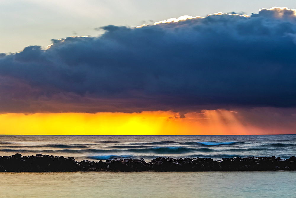 Dramatic sunrise over Lydgate Beach, breakwater and the ocean from the coast of Kauai with dark storm clouds and a distant rainfall; Kapaa, Kauai, Hawaii, United States of America