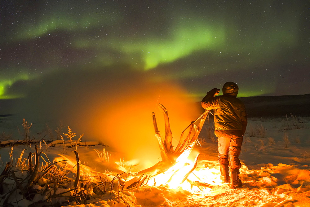 Staying warm beside a campfire on the Delta River while watching the aurora borealis on a frigid night; Alaska, United States of America