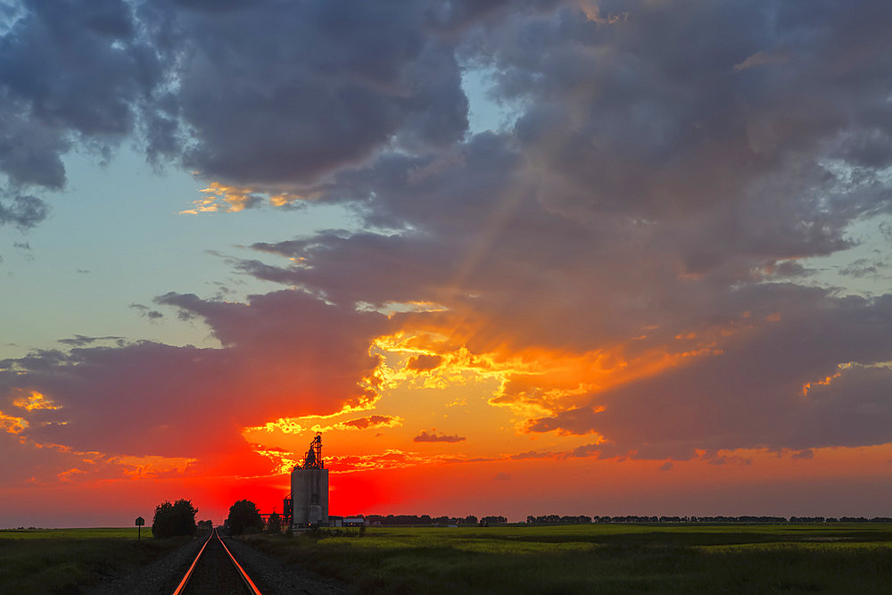 A grain storage facility at sunset with sinking sun in a glowing pink sky behind a farm; Alberta, Canada