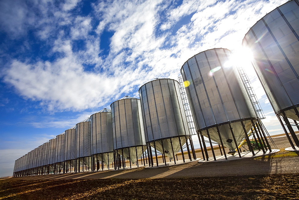 Silver metal grain storage bins in a row against a blue sky with cloud; Alberta, Canada