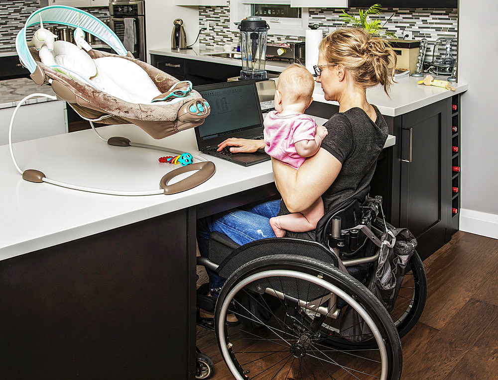 A young mom with a spinal cord injury looks after her newborn baby while working on her computer in the kitchen; Edmonton, Alberta, Canada