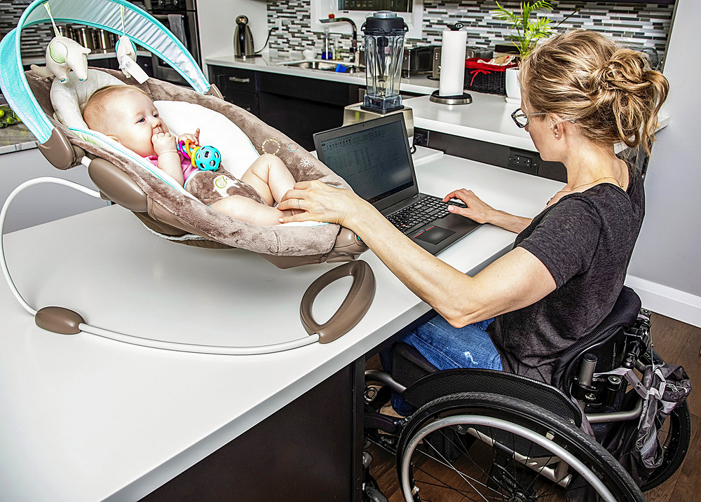 A young mom with a spinal cord injury looks after her newborn baby while working on her computer in the kitchen; Edmonton, Alberta, Canada