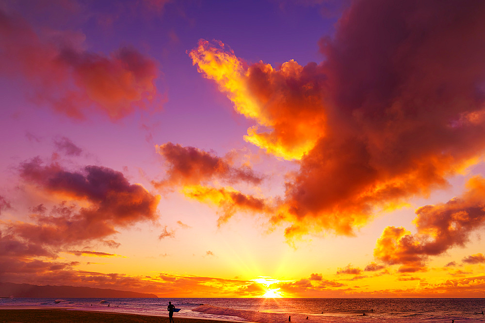 Silhouette of a surfer standing on Kelki Beach at the water's edge with dramatic glowing clouds above at sunset; Oahu, Hawaii, United States of America