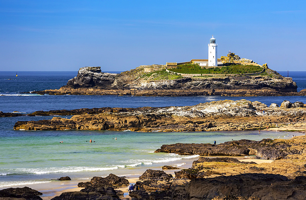 White Godrevy Lighthouse on rock formation in blue water with blue sky and rocky shoreline, Godrevy Island in St. Ives Bay; Cornwall County, England