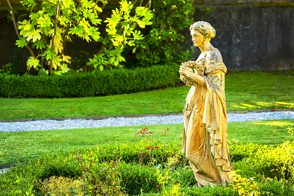 Stone female statue in garden with pebble pathway, framed by stone wall and trees; Cornwall County, England