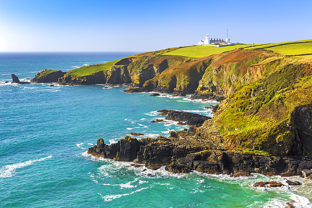 White lighthouse on top of hilly green fields framed by rocky outcrops and blue sky; Cornwall County, England