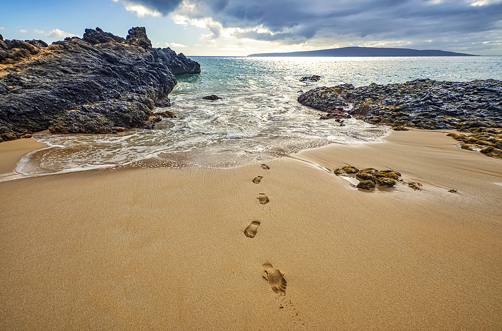 Footprints on the sand lead to the shore from the water's edge, with rugged lava rock lining the coastline; Kihei, Maui, Hawaii, United States of America